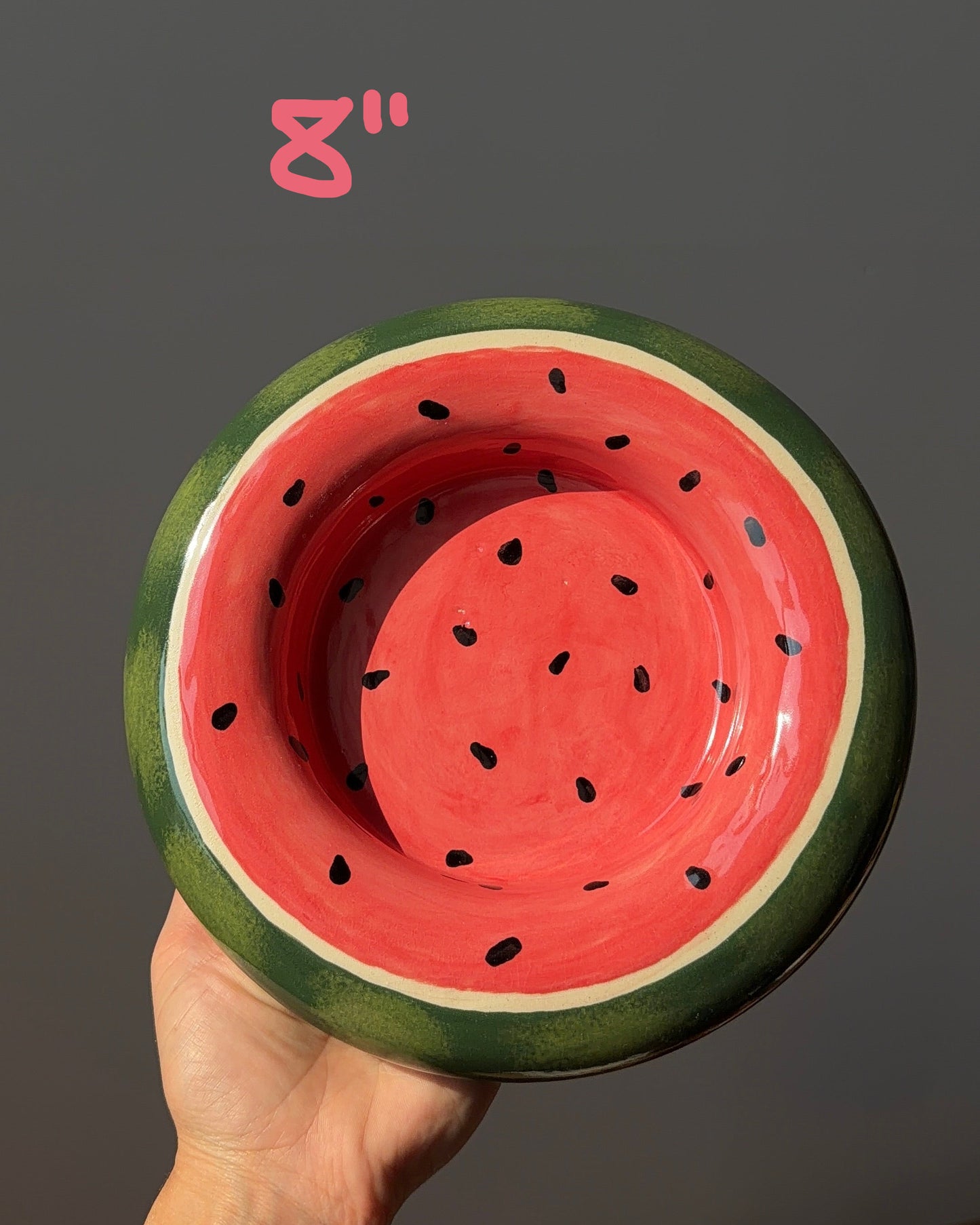A hand holding a ceramic bowl designed to look like a watermelon slice, featuring a red inner section with black seeds, a white rind line, and a dark green outer rind, set against a dark gray background.