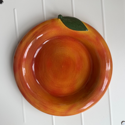 An orange-shaped ceramic bowl with a green leaf on top, displayed on a white plastic tray over a wooden floor.