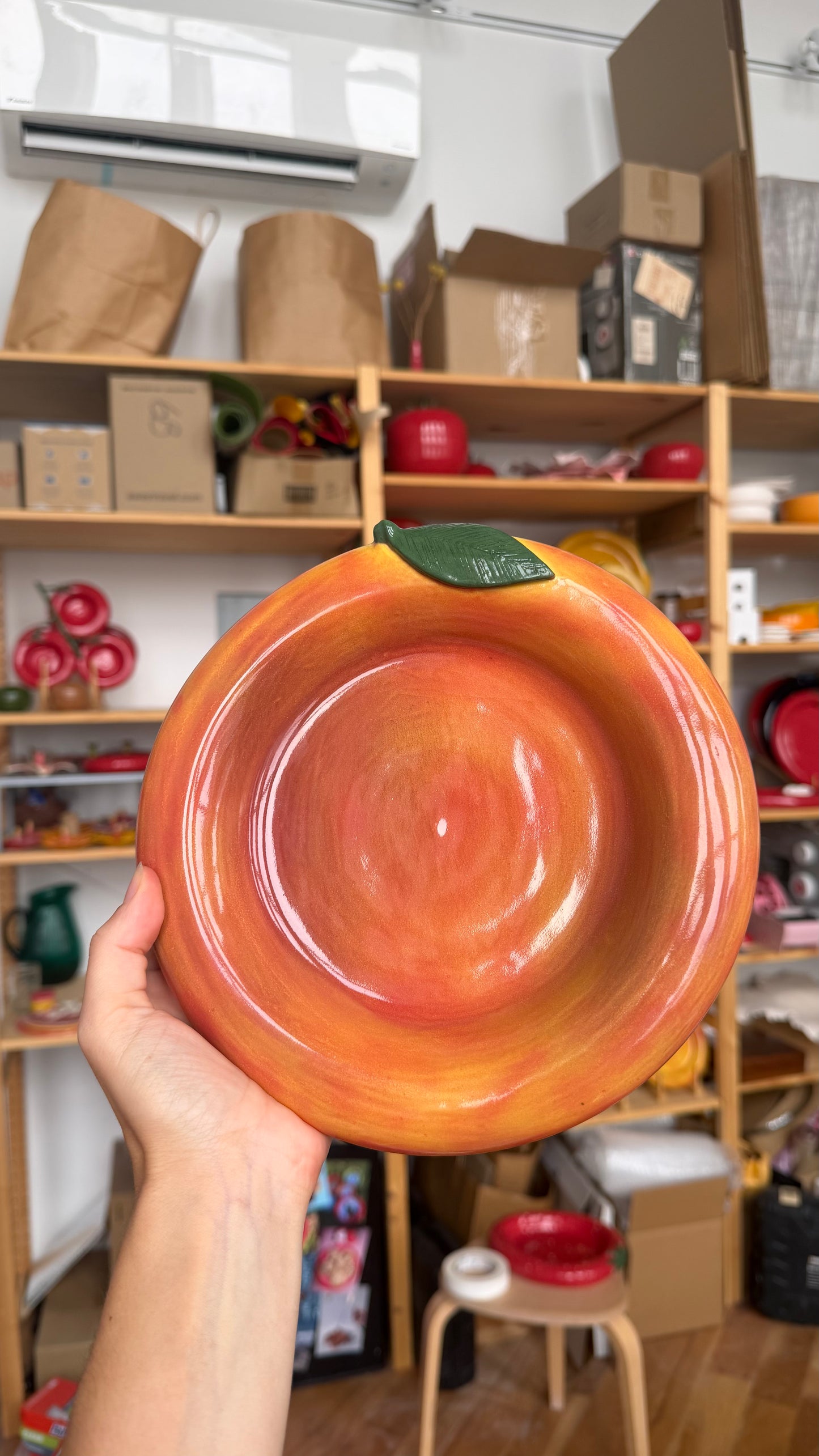 A hand holds a large, glossy ceramic bowl shaped like a peach or orange, featuring a vibrant orange and yellow gradient with a green leaf on top, set against a cluttered background of wooden shelves filled with boxes and other items.
