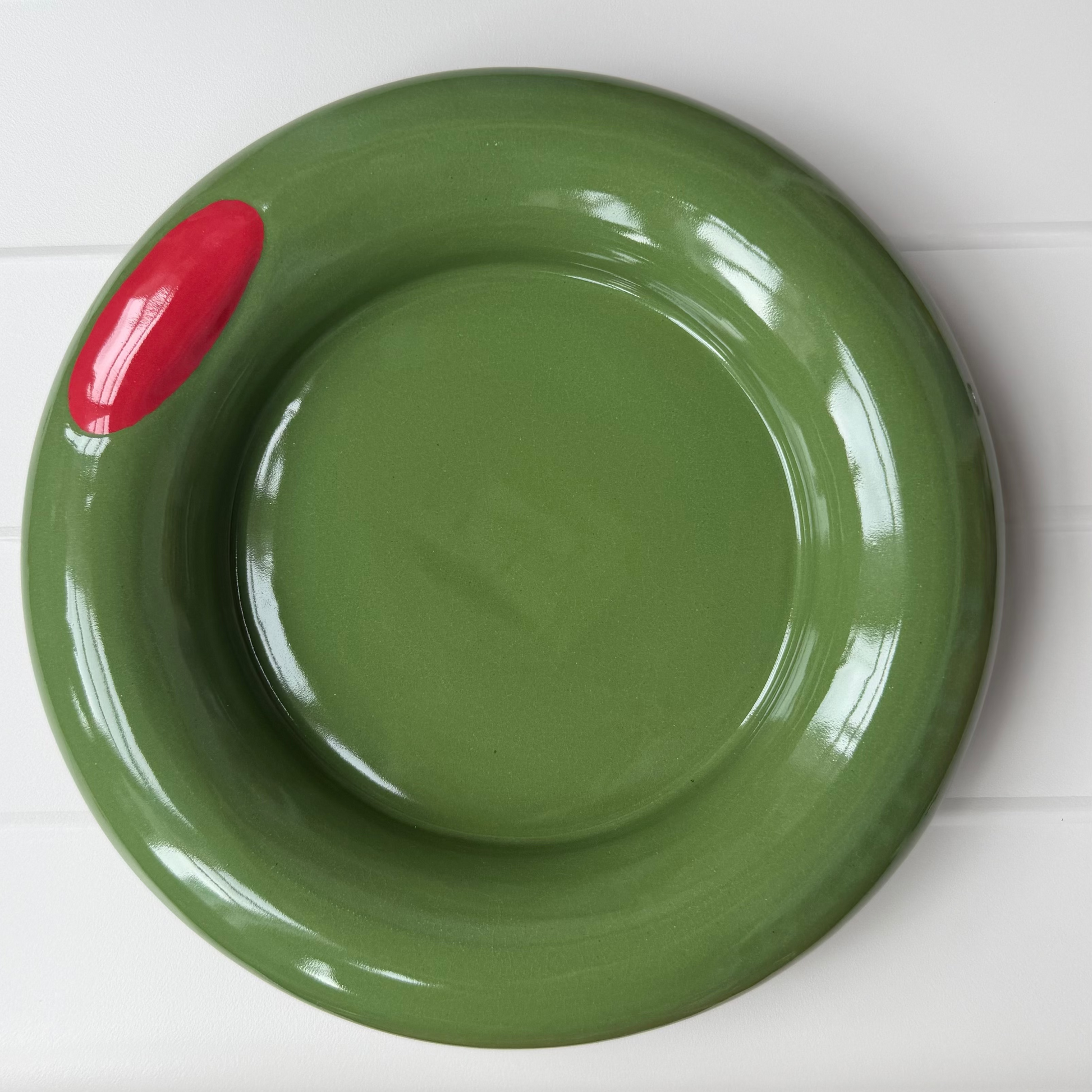 A glossy olive green ceramic plate with a small red accent on the left side, photographed from above on a white table.