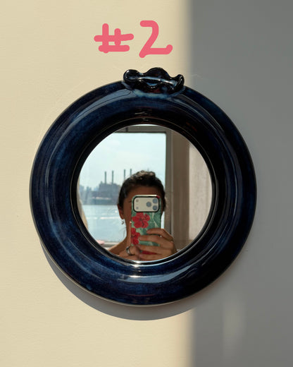 A round ceramic blueberry wall mirror with a thick, glossy dark blue ceramic frame, mounted on a beige wall with a strong diagonal shadow. The mirror reflects a person taking a selfie.
