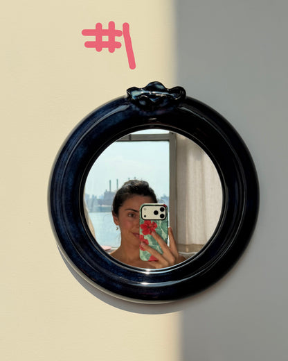 A round blueberry ceramic wall mirror with a glossy dark navy blue frame, mounted on a beige wall. The mirror reflects a person taking a selfie with a phone.