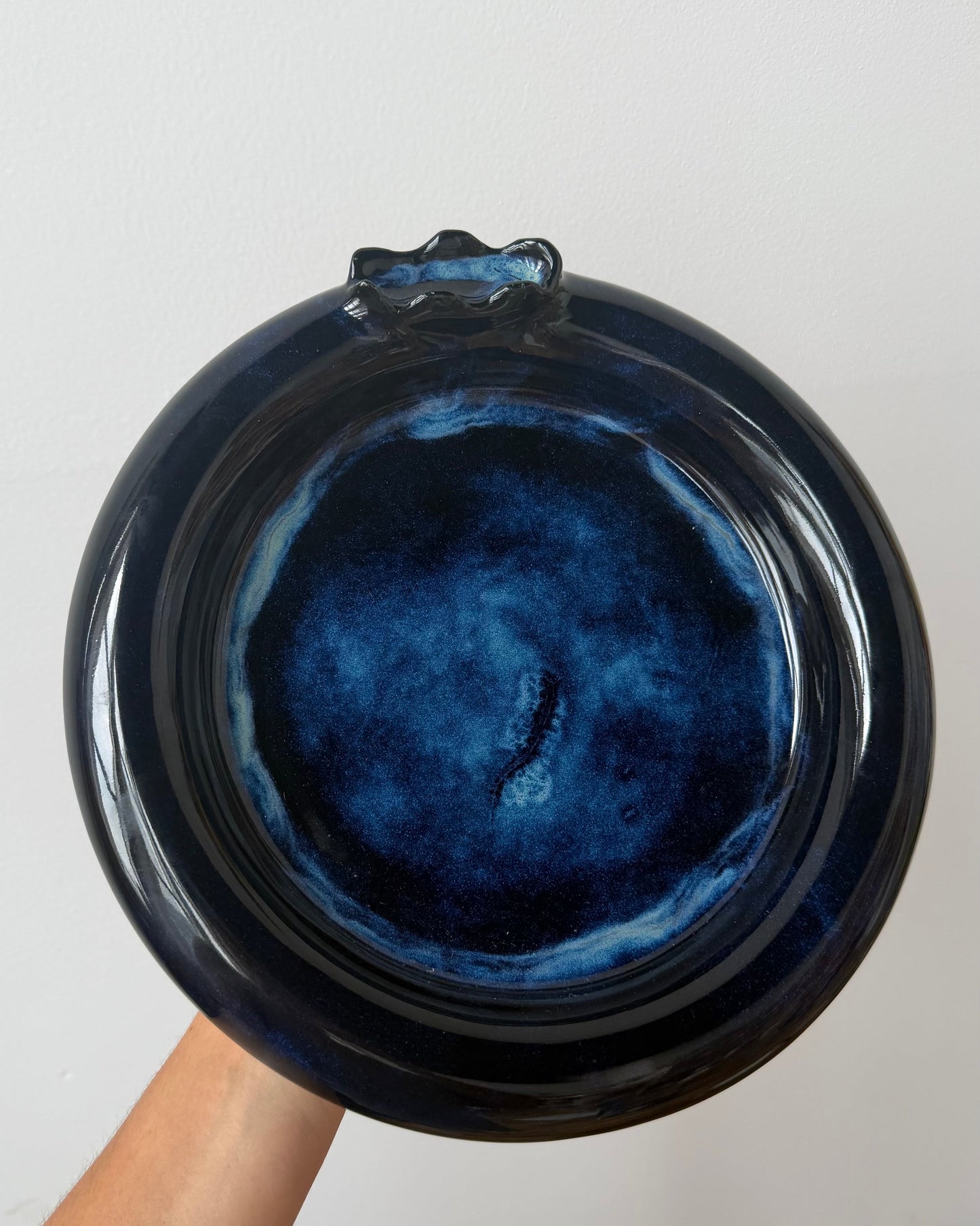 A hand holding a round glossy ceramic plate or dish with a deep blue and black glaze, photographed against a plain light background.