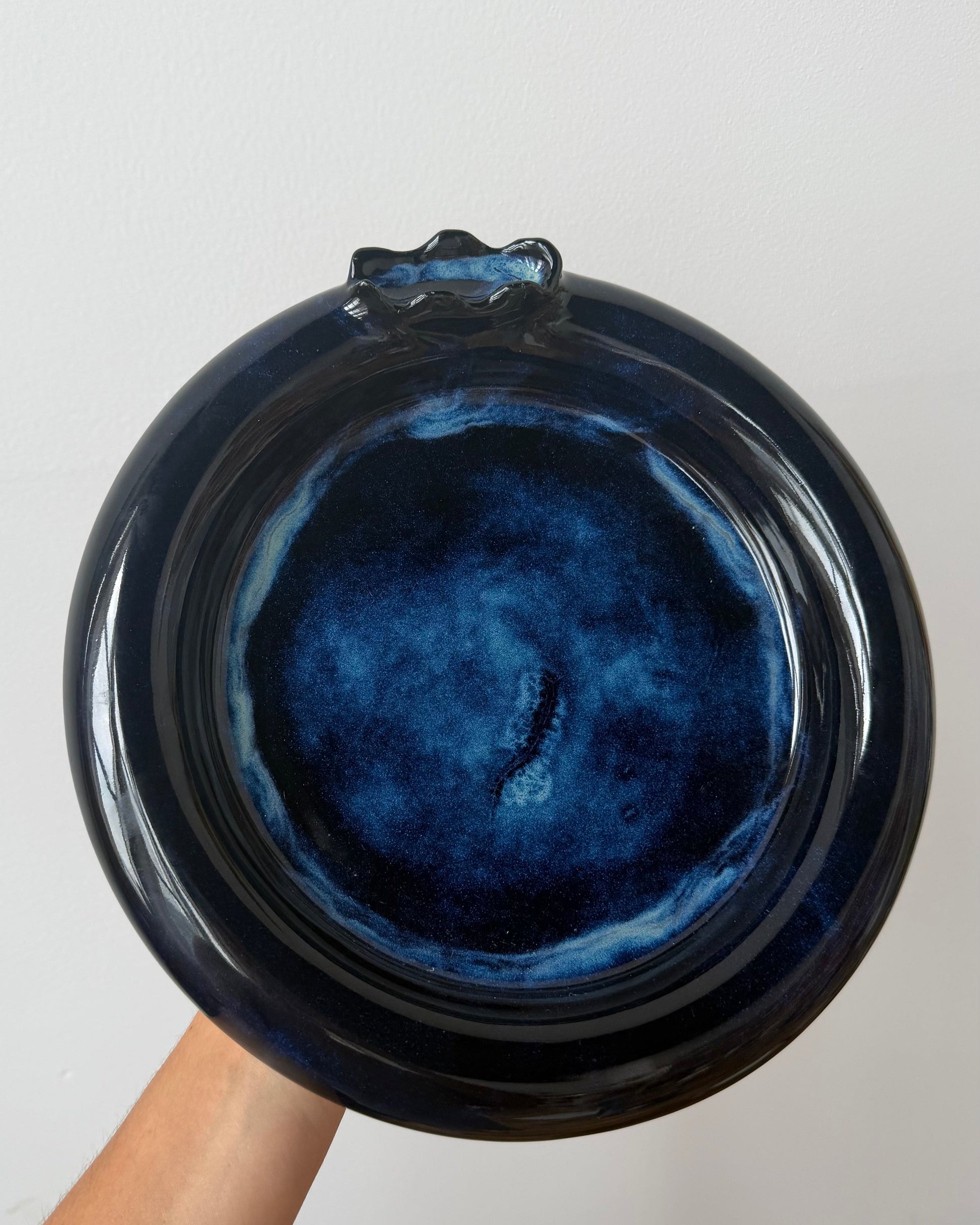 A hand holding a round glossy ceramic plate or dish with a deep blue and black glaze, photographed against a plain light background.