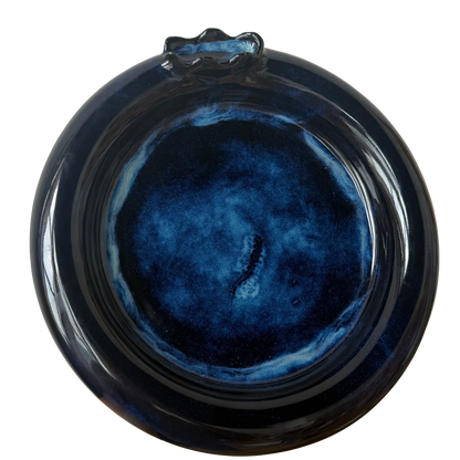 A round ceramic plate with a glossy dark blue and black marbled glaze, featuring a small decorative protruding handle at the top, shown against a plain white background.