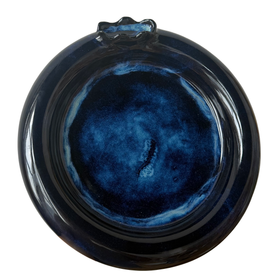 A round ceramic plate with a glossy dark blue and black marbled glaze, featuring a small decorative protruding handle at the top, shown against a plain white background.