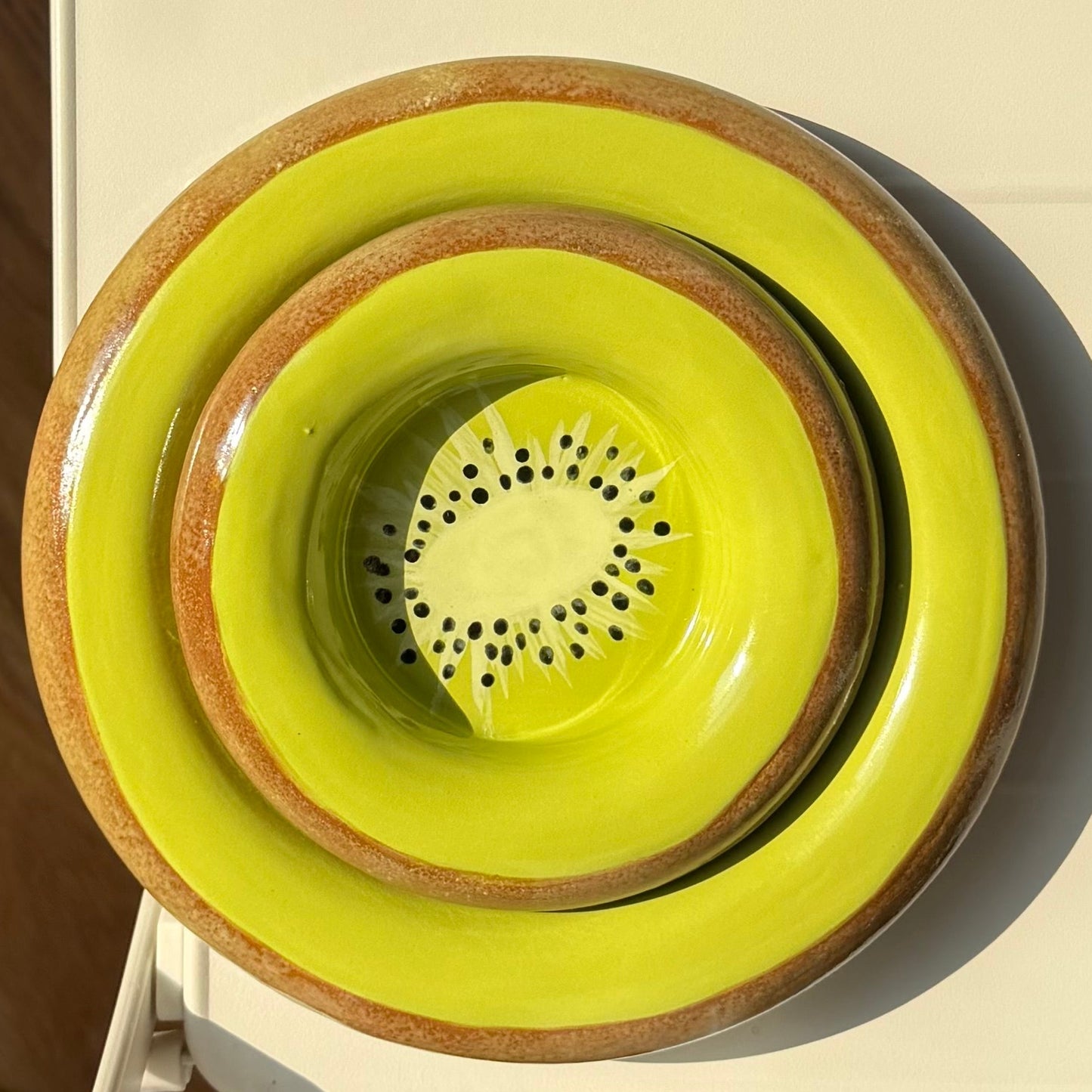 A stack of three ceramic bowls in bright yellow-green and brown, viewed from above on a white table with a wooden floor and part of a person's legs and shoes visible.