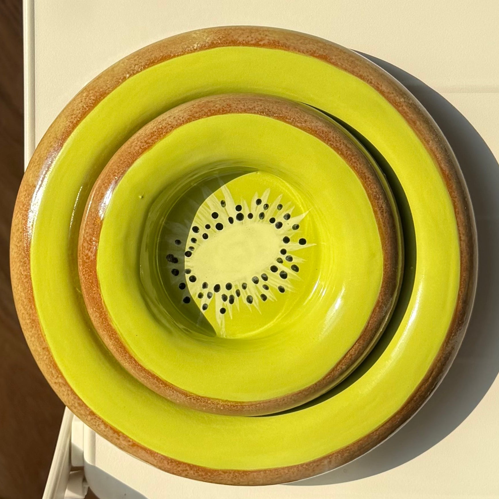 A stack of three ceramic bowls in bright yellow-green and brown, viewed from above on a white table with a wooden floor and part of a person's legs and shoes visible.