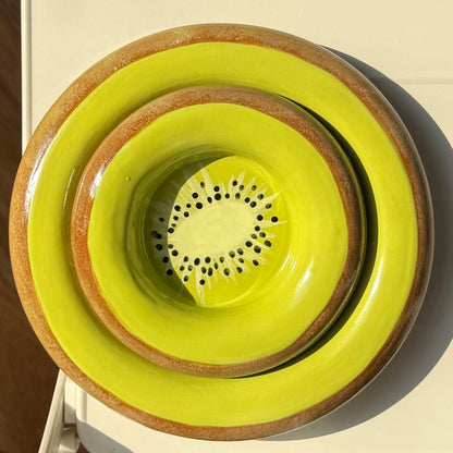A stack of three ceramic bowls in bright yellow-green and brown, viewed from above on a white table with a wooden floor and part of a person's legs and shoes visible.