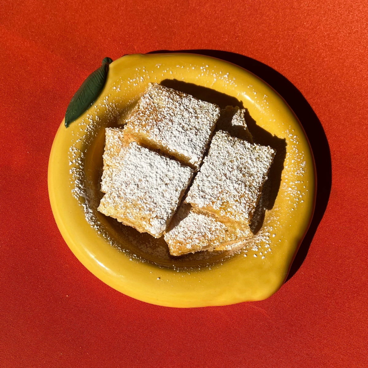Three square lemon bars with powdered sugar on a yellow lemon bubble bowl against a red background