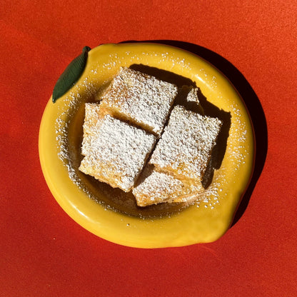 Three square lemon bars with powdered sugar on a yellow lemon bubble bowl against a red background