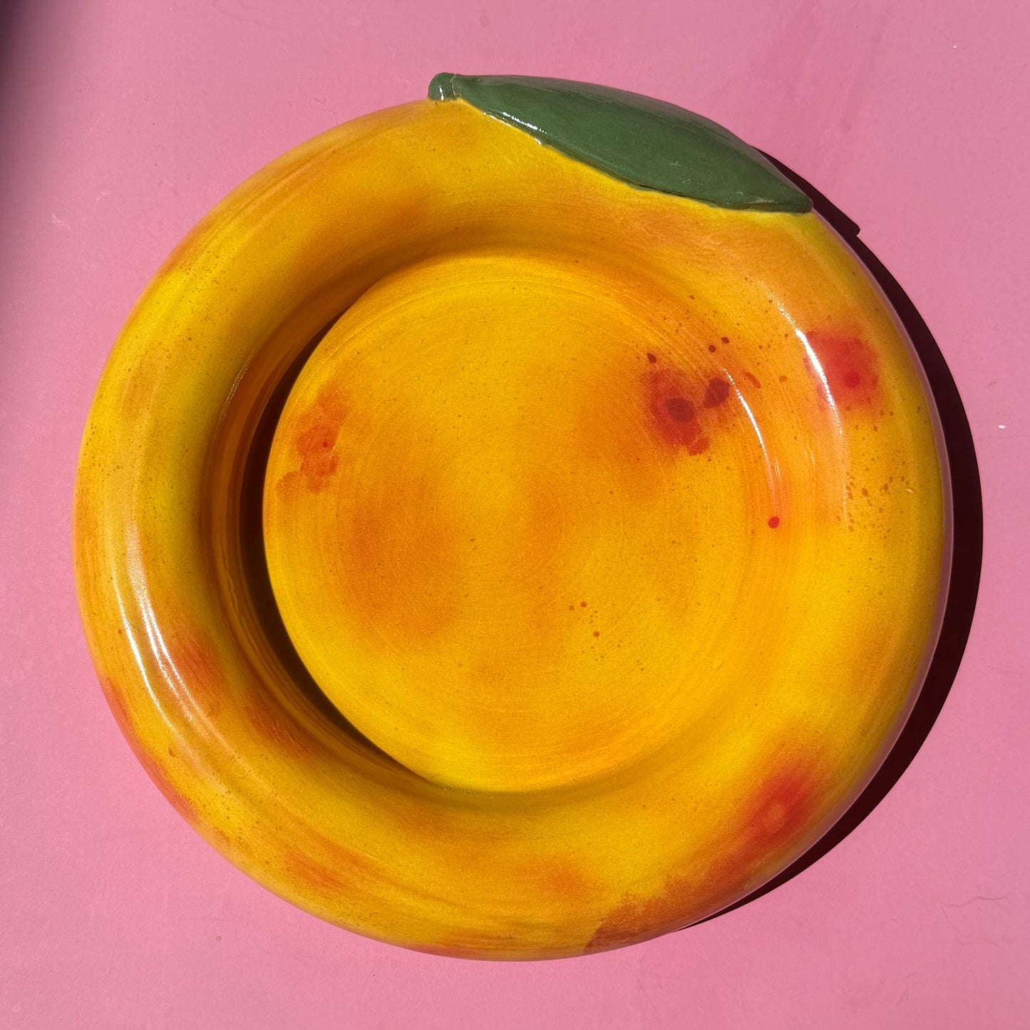 A ceramic bowl painted to look like a peach, featuring a yellow and orange body with red spots and a green leaf accent, set against a pink background.