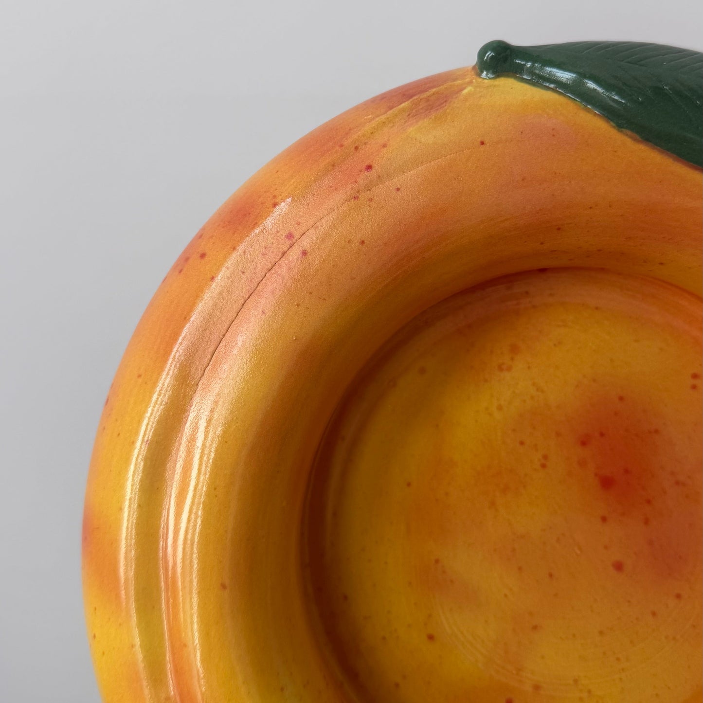 A close-up of a ceramic bowl shaped like a peach, showing its glossy yellow and orange surface with subtle red speckles and a dark green stem, held against a plain light gray background.