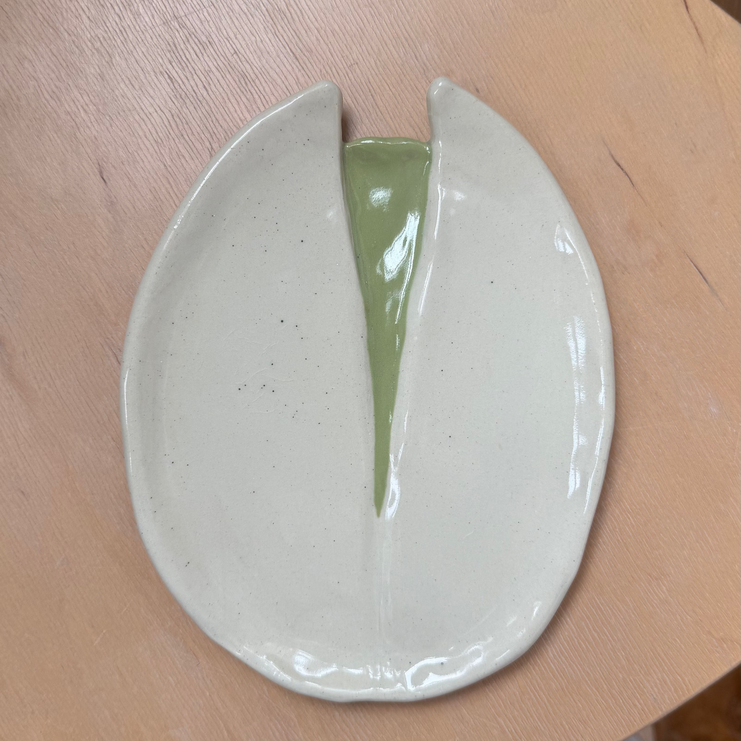 A handmade ceramic dish with a glossy off-white exterior and a light green glazed center, shown resting on a light wood surface.