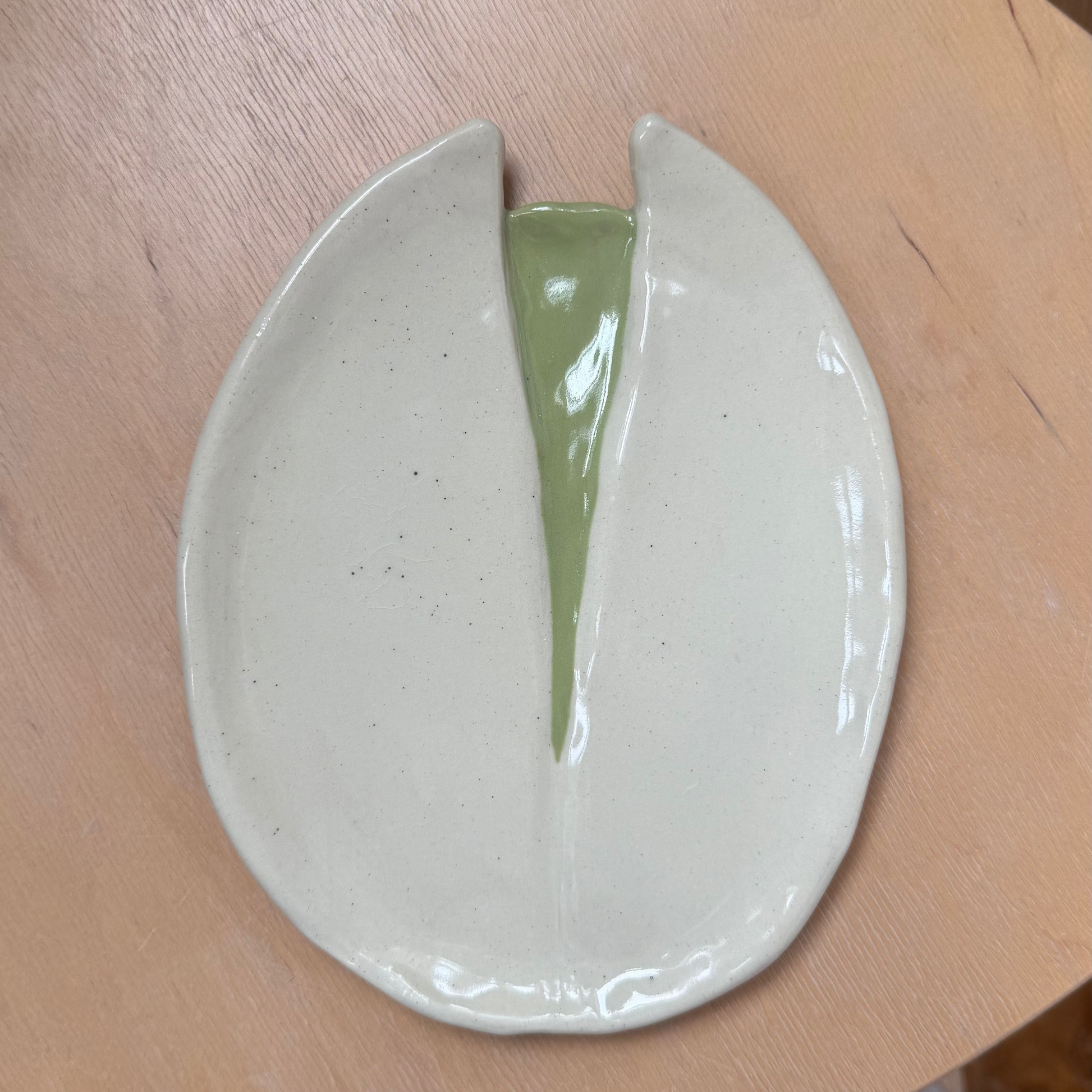 A handmade ceramic dish with a glossy off-white exterior and a light green glazed center, shown resting on a light wood surface.
