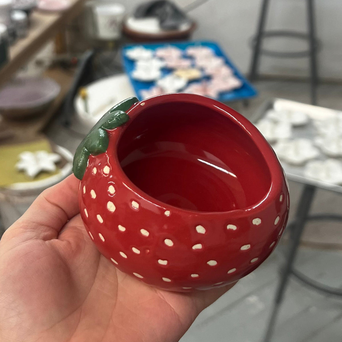 A hand holding a small red ceramic bowl shaped like a strawberry, featuring white polka dots and a green leaf detail, in a workshop setting.
