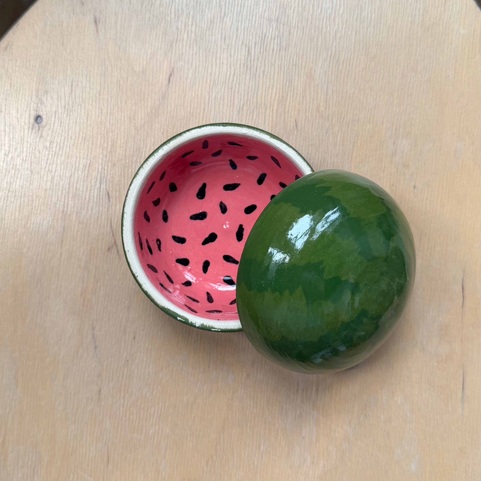 A small decorative bowl shaped like a watermelon half, with a green rind-covered sphere lid resting on top and the inside of the bowl showing a pink pattern with black seed details, placed on a light wood table.